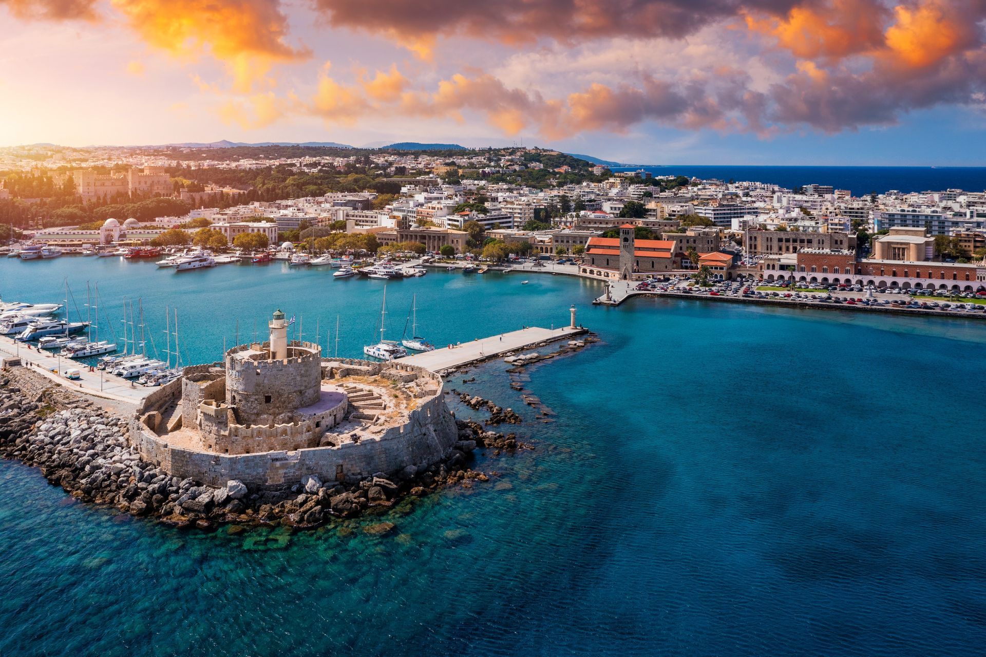 Aerial view of a coastal harbor with turquoise water, stone breakwater, and a sunset-lit seaside town.