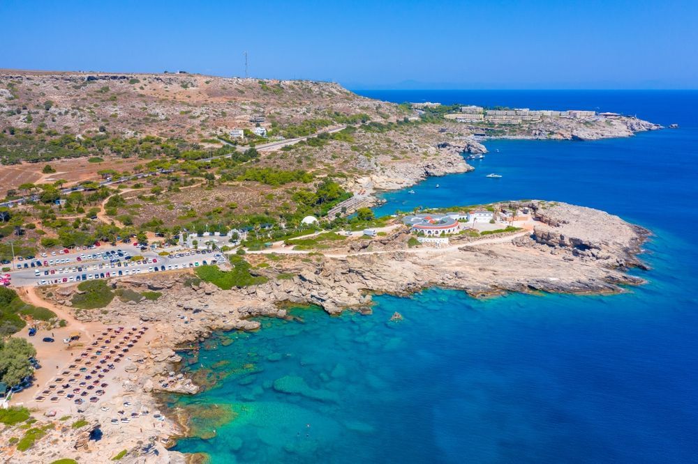 Aerial view of a rocky coastline with turquoise water, sandy beach, and white buildings near the shore