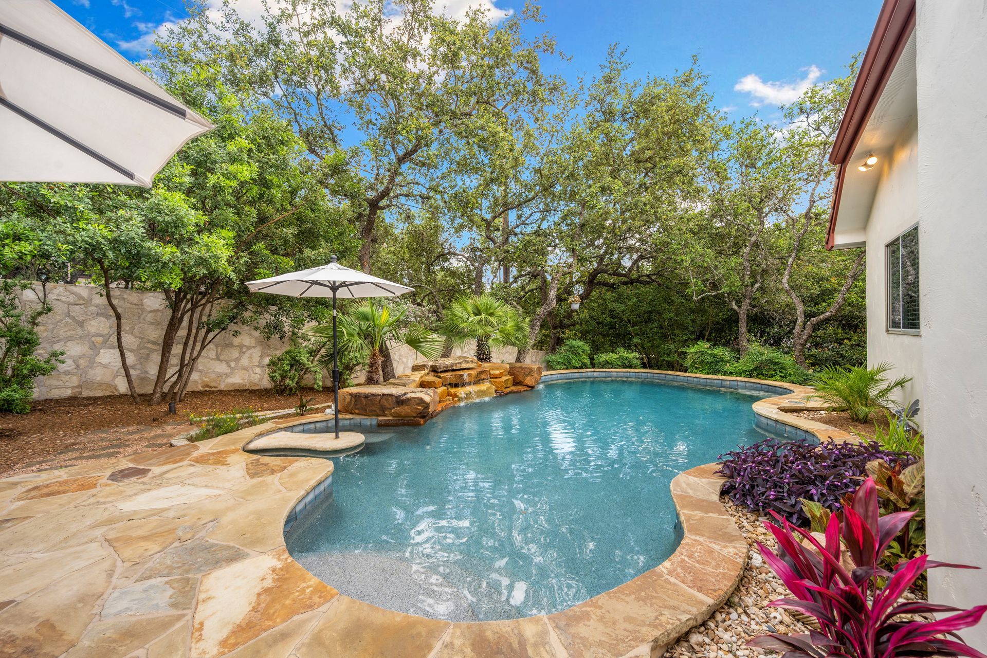 Backyard pool with stone patio, lounge chairs, umbrellas, and lush greenery under a blue sky