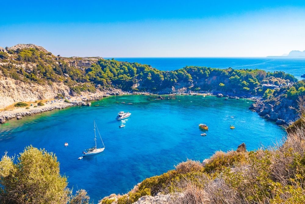 Turquoise bay with sailboats, rocky cliffs, and dense greenery under a clear blue sky