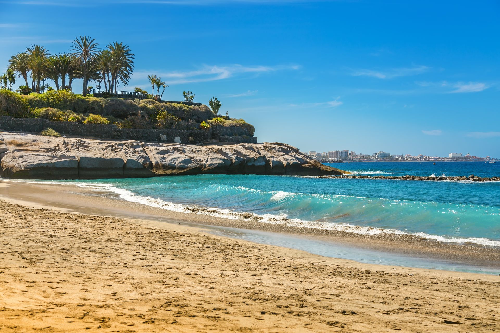 Sunny beach with turquoise water, sandy shore, rocky cliffs, and palm trees under a clear blue sky
