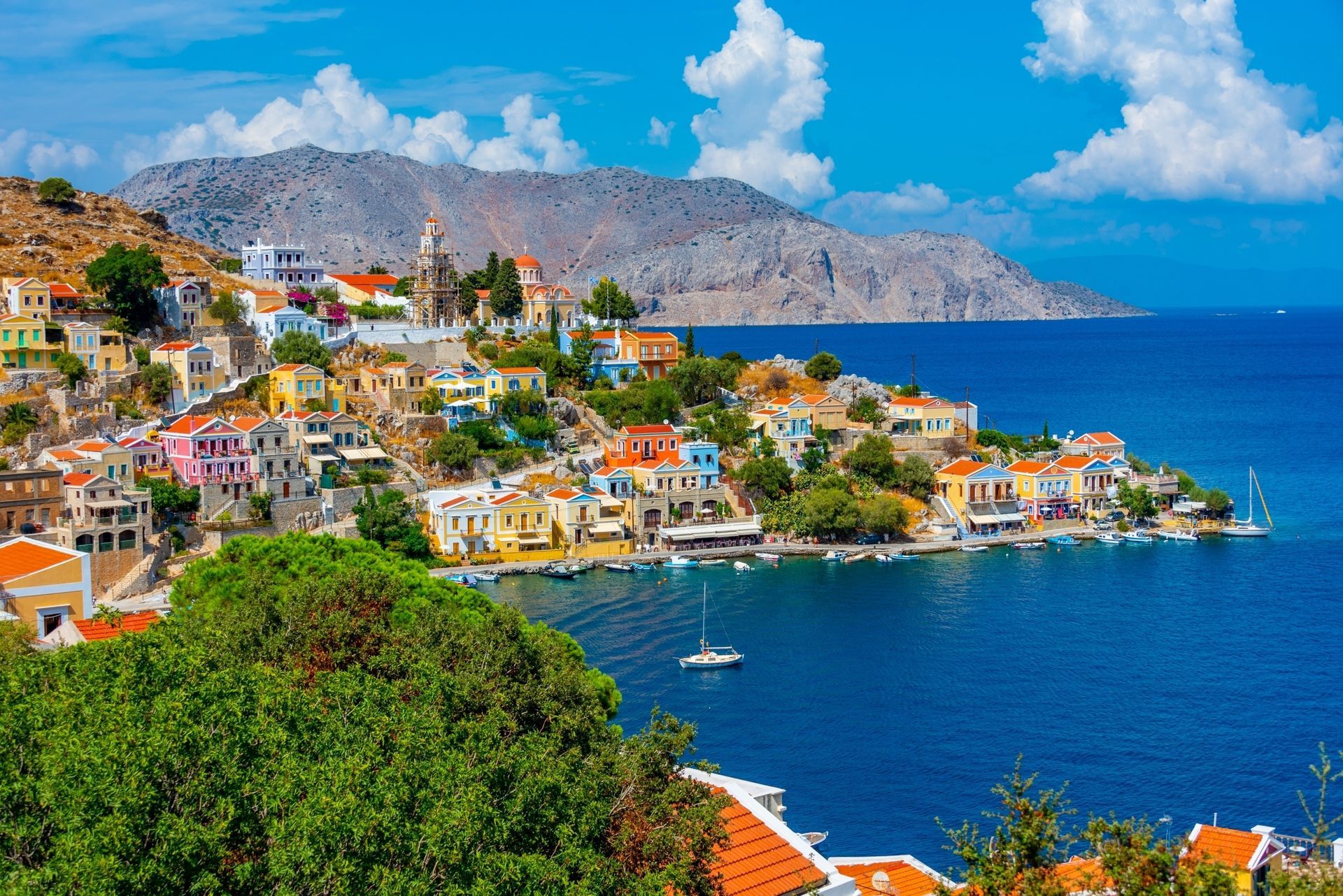 Colorful hillside seaside village with red-roofed houses, blue water, and mountains under a bright sky