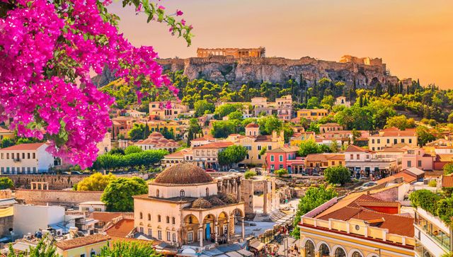Blooming pink bougainvillea frames a hillside cityscape with stone buildings under a warm sunset sky.