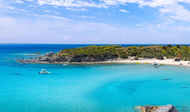 Turquoise bay with white sandy beach, rocky coastline, and a few boats under a clear blue sky