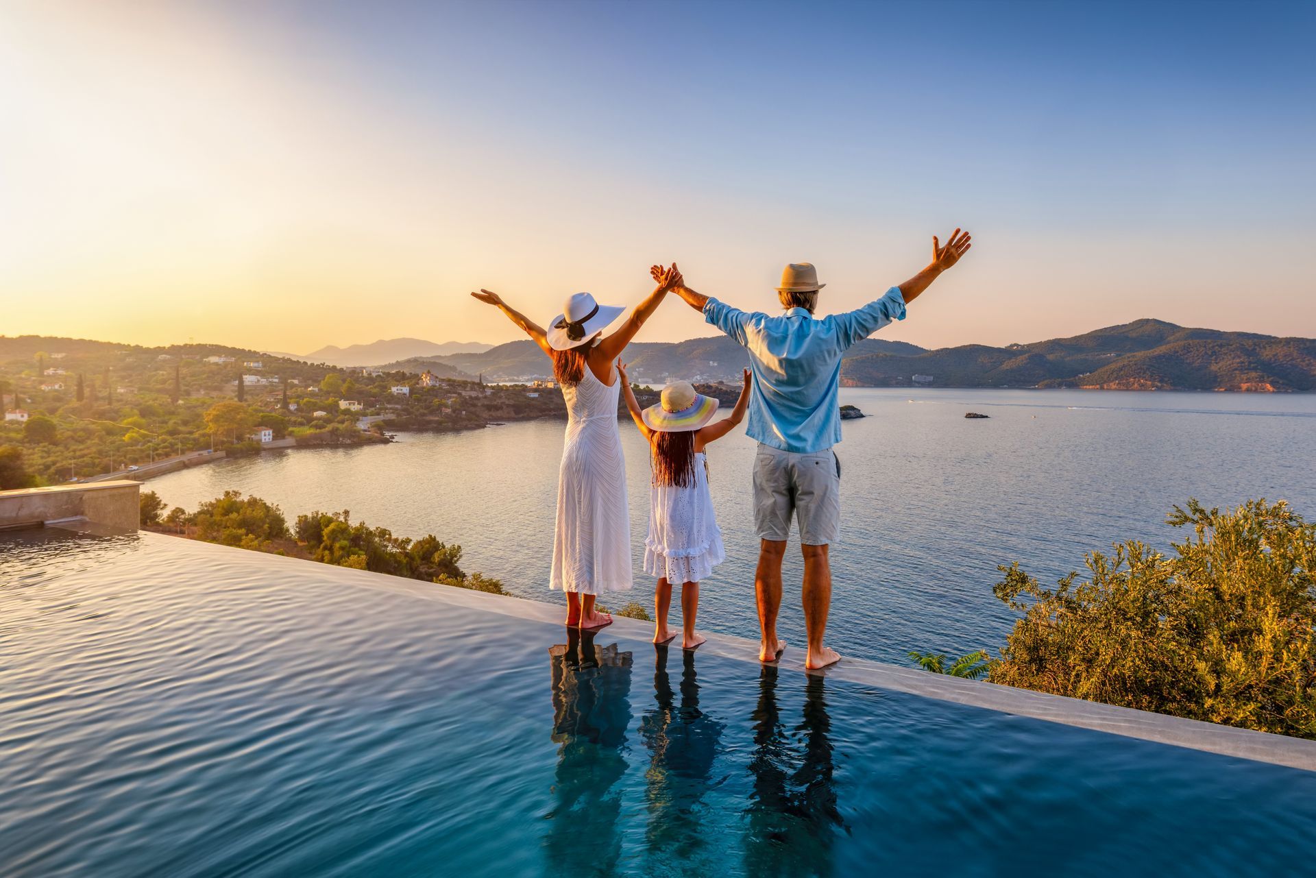 Three people stand on an infinity pool edge, arms raised, overlooking a sunset bay and hills.