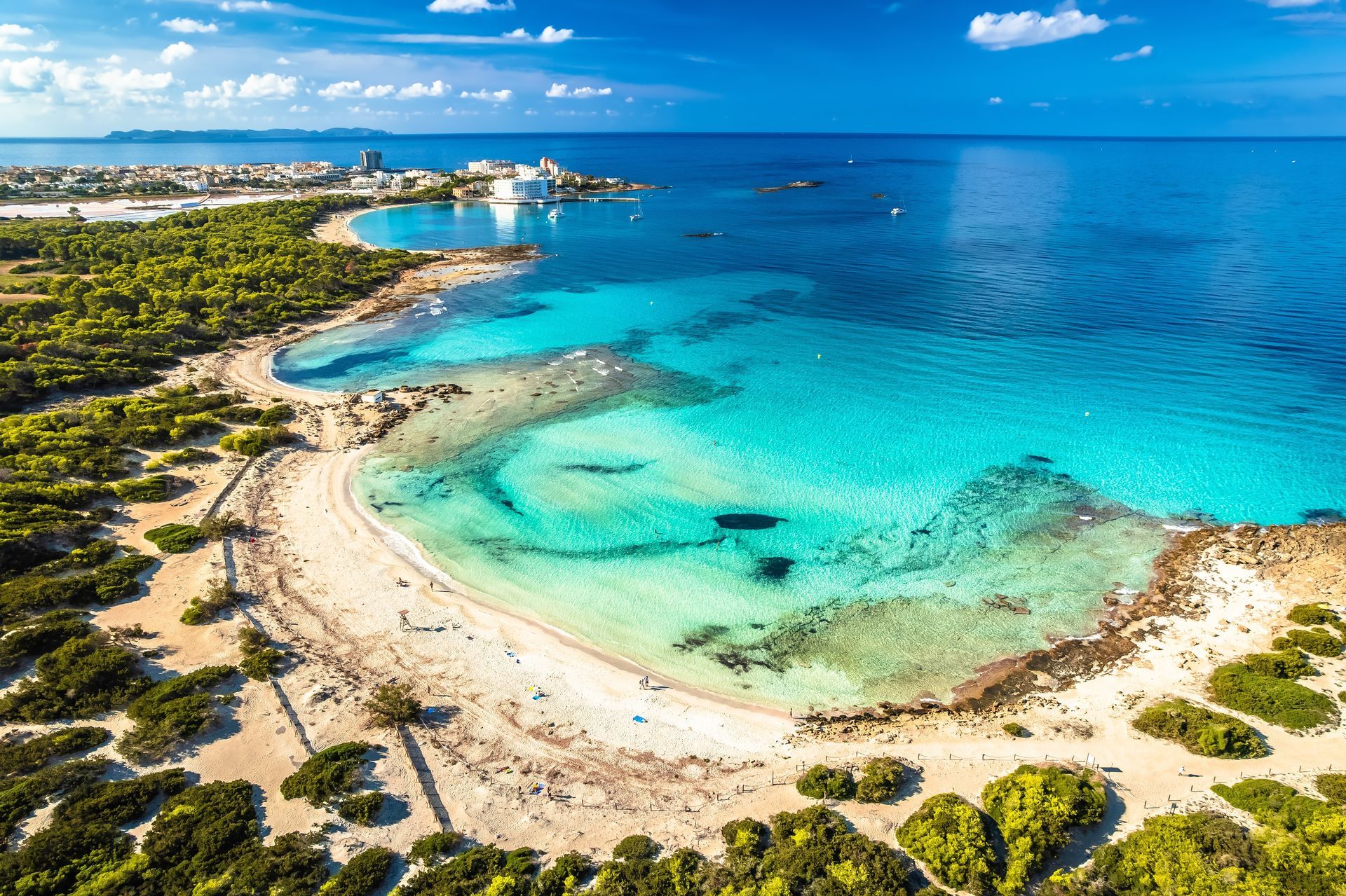 Aerial view of a tropical beach with turquoise water, sandy shore, and green coastline under a bright blue sky