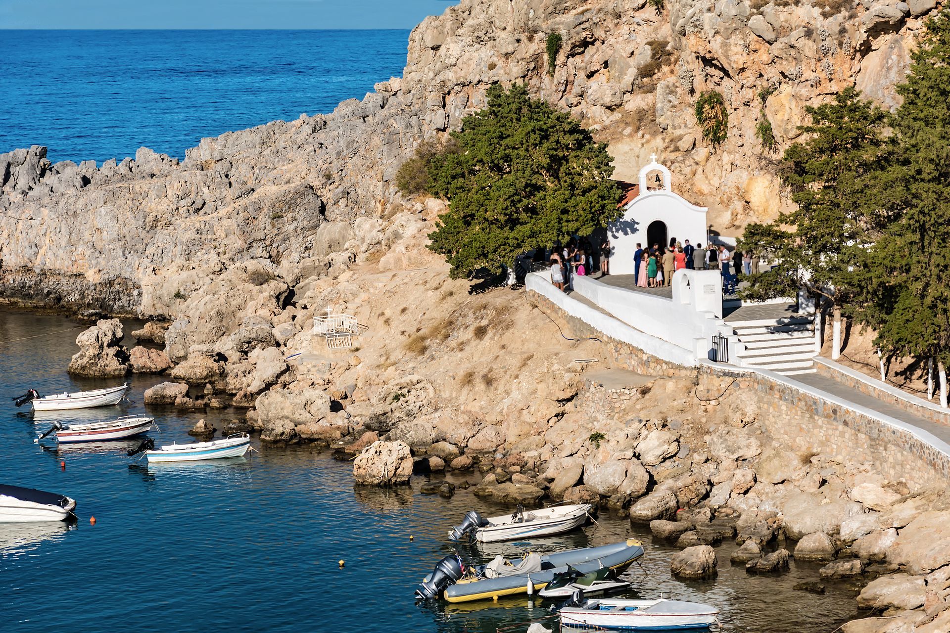 Small white chapel on rocky seaside cliff with people, blue water, and boats below