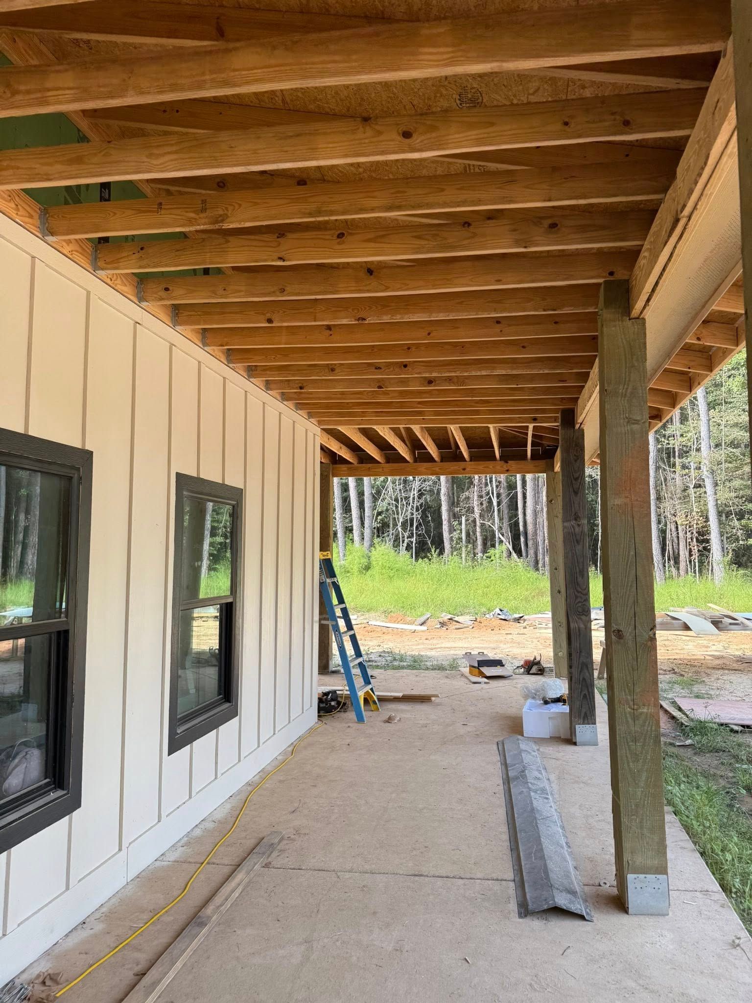 Covered porch under construction beside a house, with exposed beams, white siding, and a ladder.