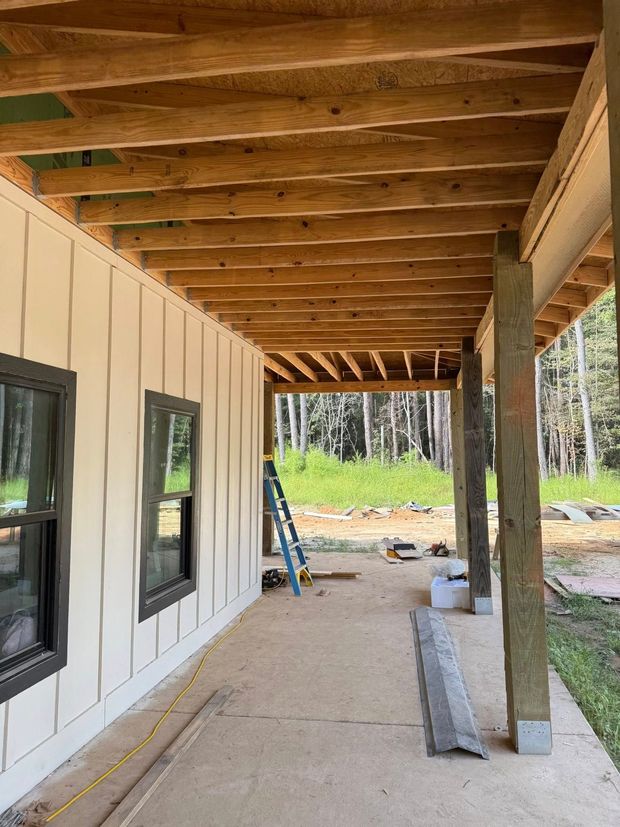 Covered porch under construction beside a house, with exposed beams, white siding, and a ladder.