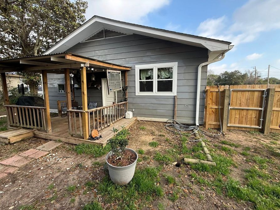 Gray house exterior with small wooden porch, fenced yard, and potted plant in the foreground