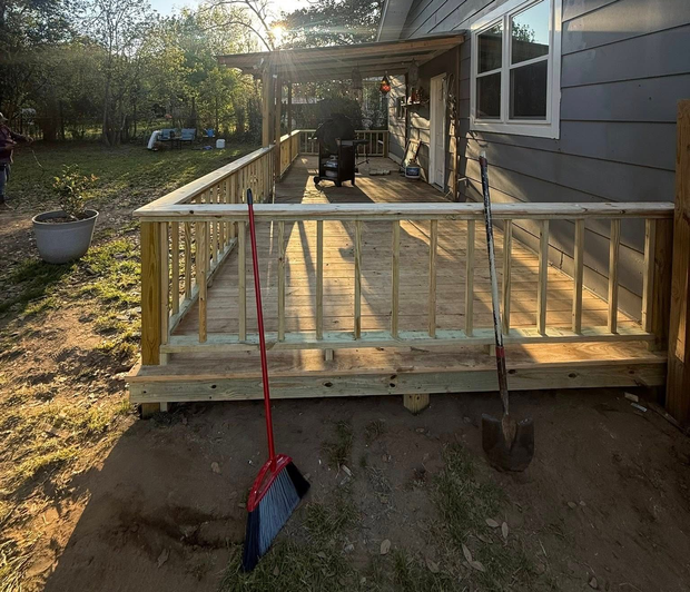 Wooden porch deck beside a gray house, with a red broom leaning against the railing.
