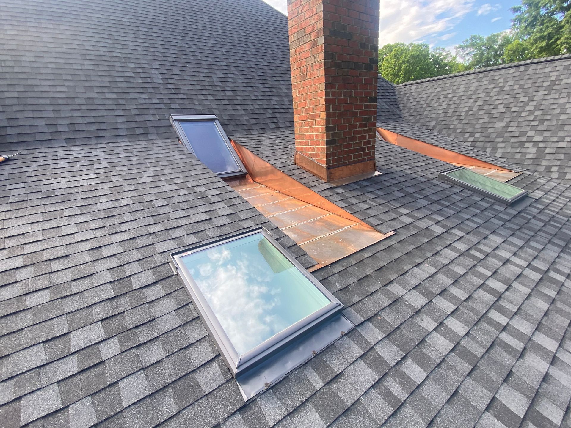 A sloped shingle roof with a brick chimney, two skylights, and exposed copper flashing.