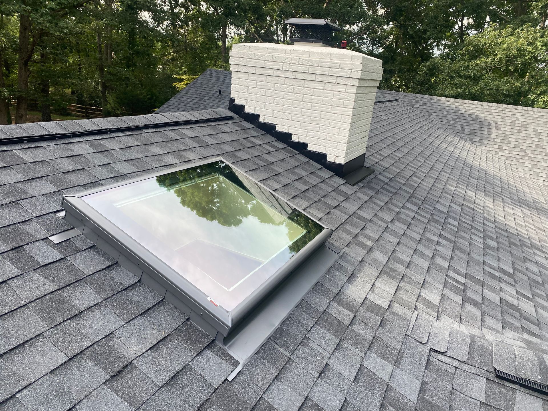A skylight installed on a dark shingled roof next to a white brick chimney.