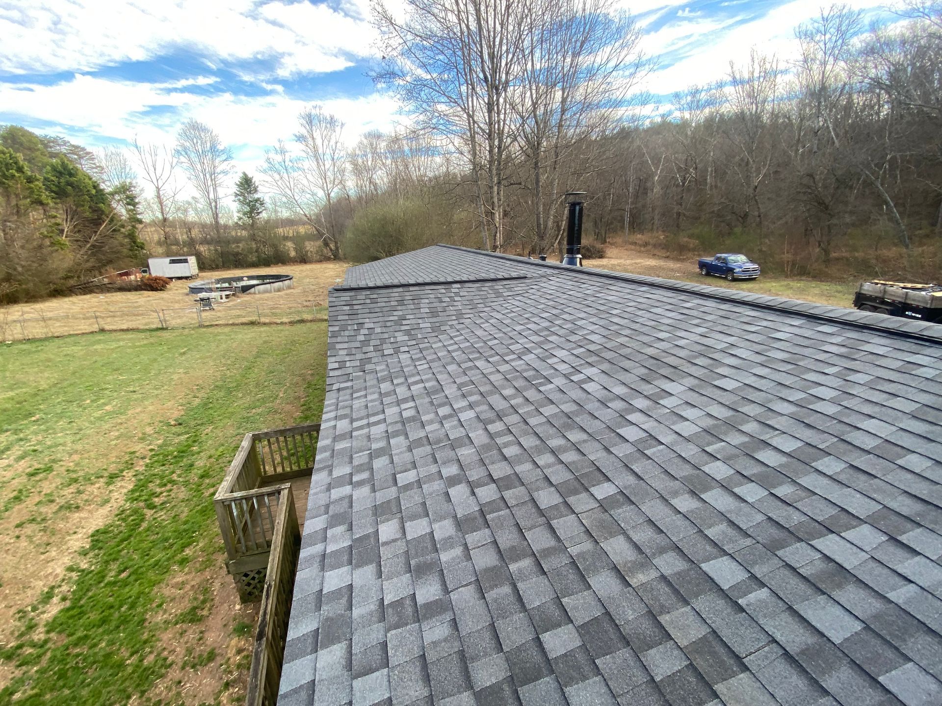 A view looking down over a gray shingled roof, showing a valley junction and a backyard with a wooden deck and trees.