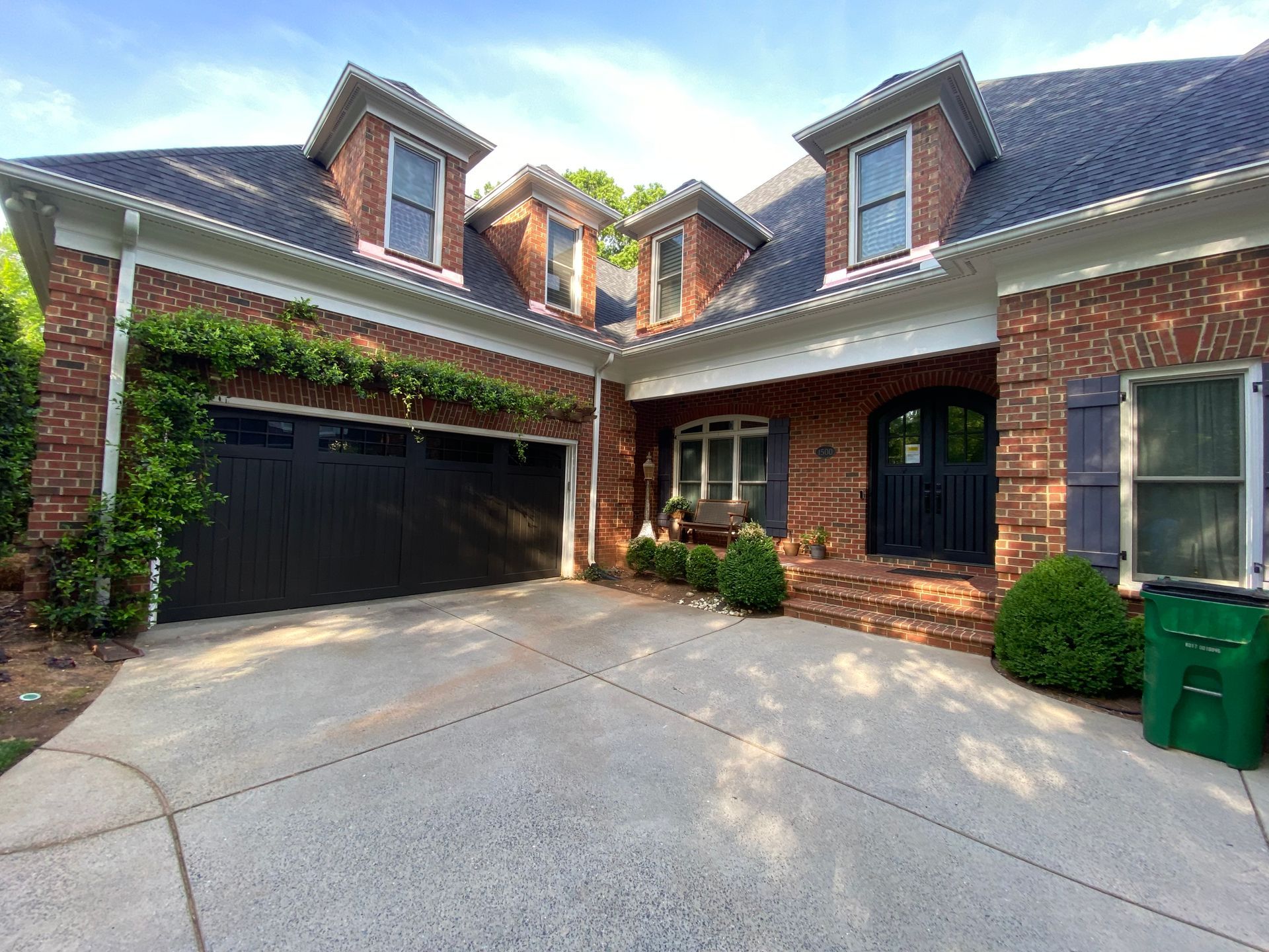 A brick house with a dark garage door, a front entrance with a black door, dormer windows, and a concrete driveway.