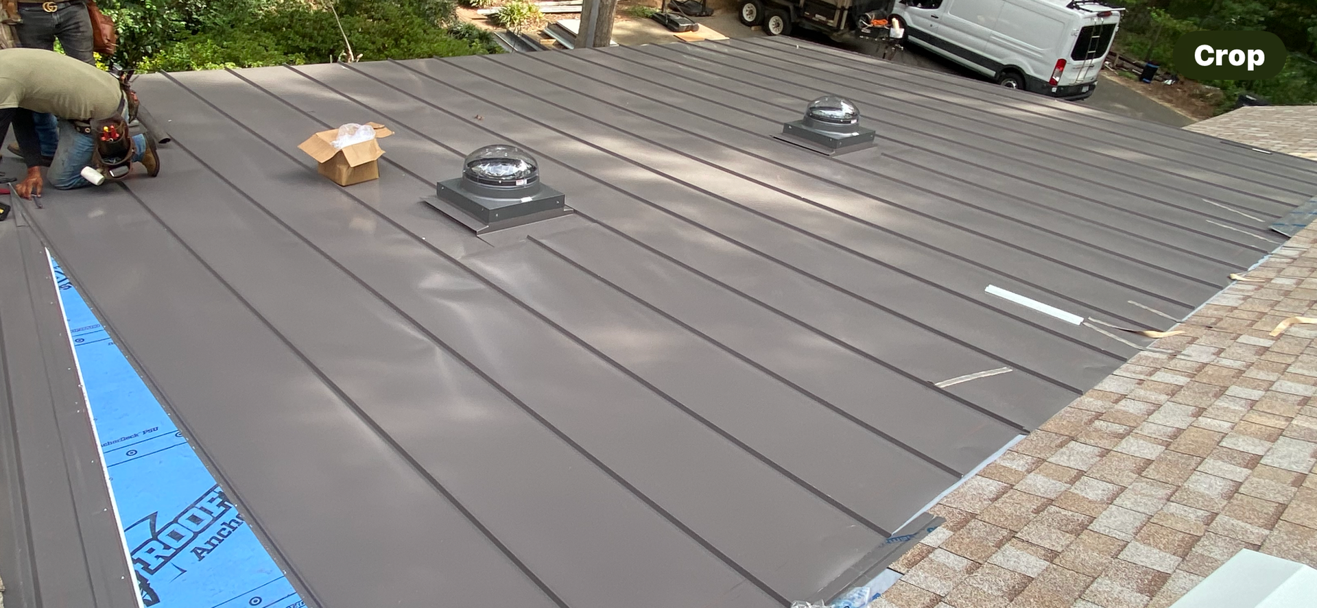 Workers installing a gray metal roof with raised seams and two ventilation pipes on a sunny day.