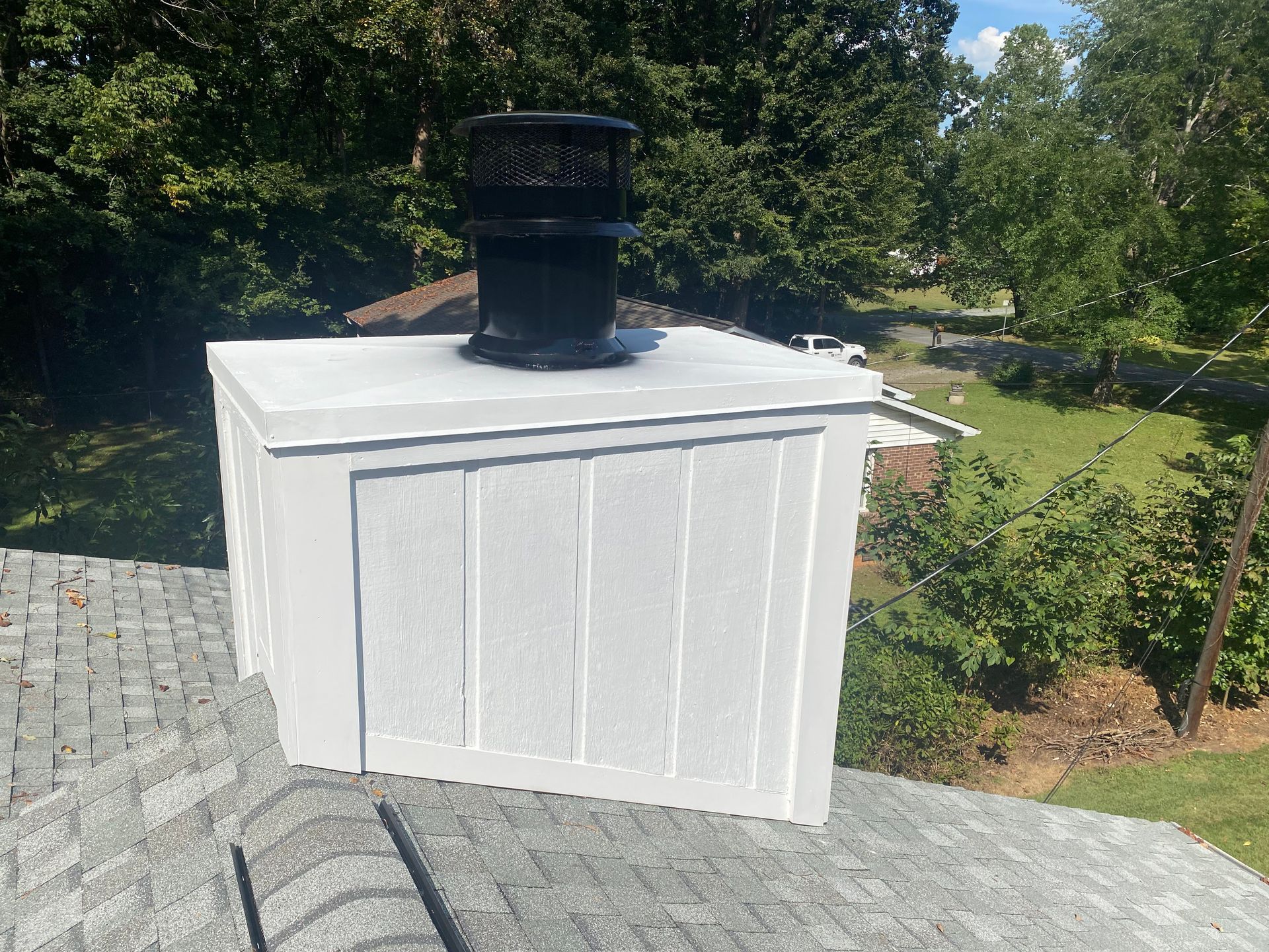 A white, rectangular chimney chase cover topped with a black metal flue vent sits on a shingled roof amid green trees.