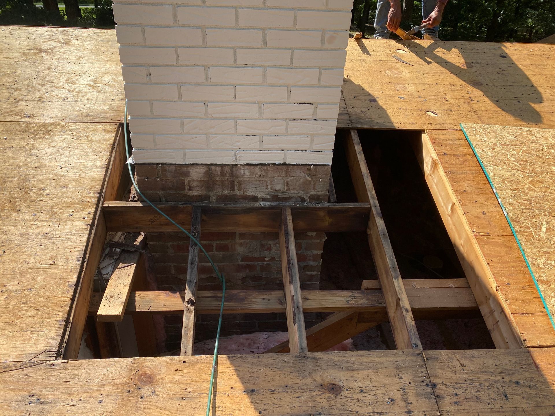 A white brick chimney rises through a roof opening, showing wooden rafters and plywood decking during a renovation.
