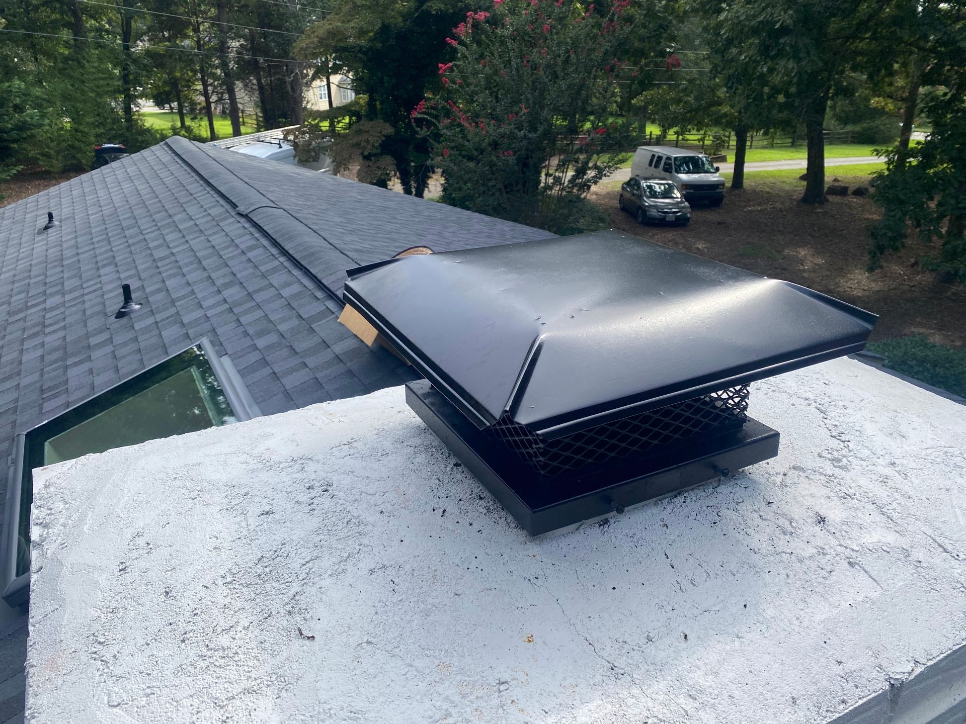A black chimney cap sits on a white concrete chimney crown on a shingled roof, with trees and a parked car in the distance.