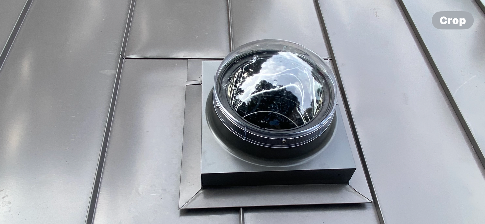 A solar tube skylight with a clear, dome-shaped cover installed on a grey metal standing-seam roof.