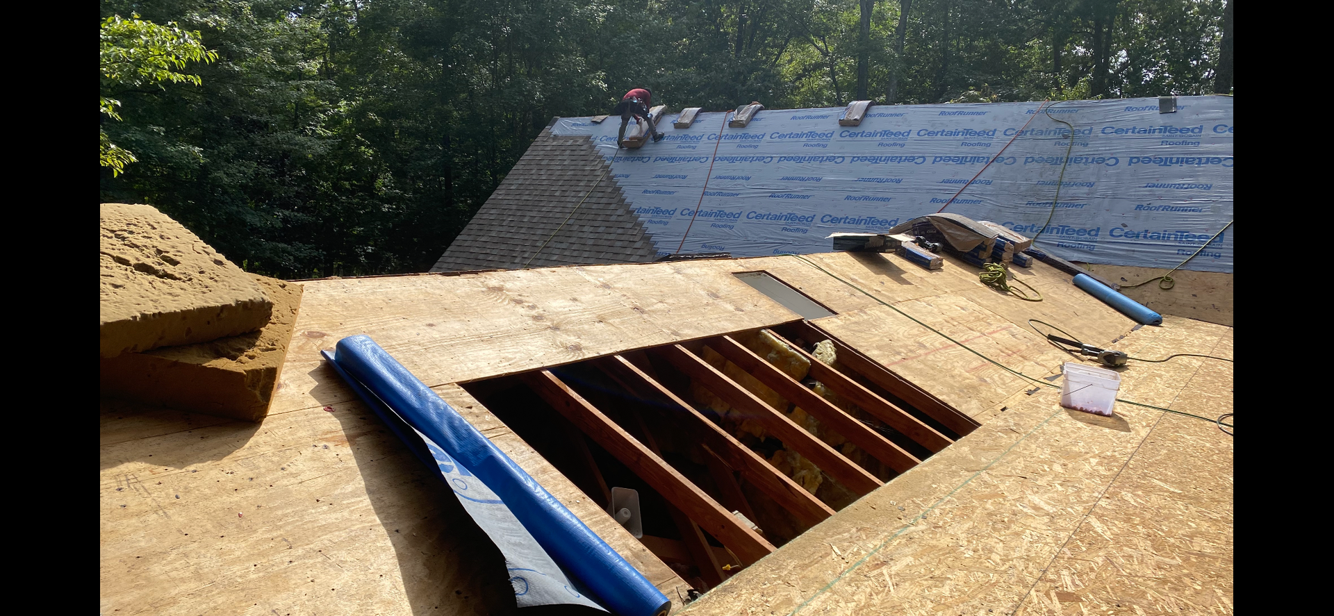 A construction worker on a roof, featuring a large rectangular opening exposing the wooden rafters below.