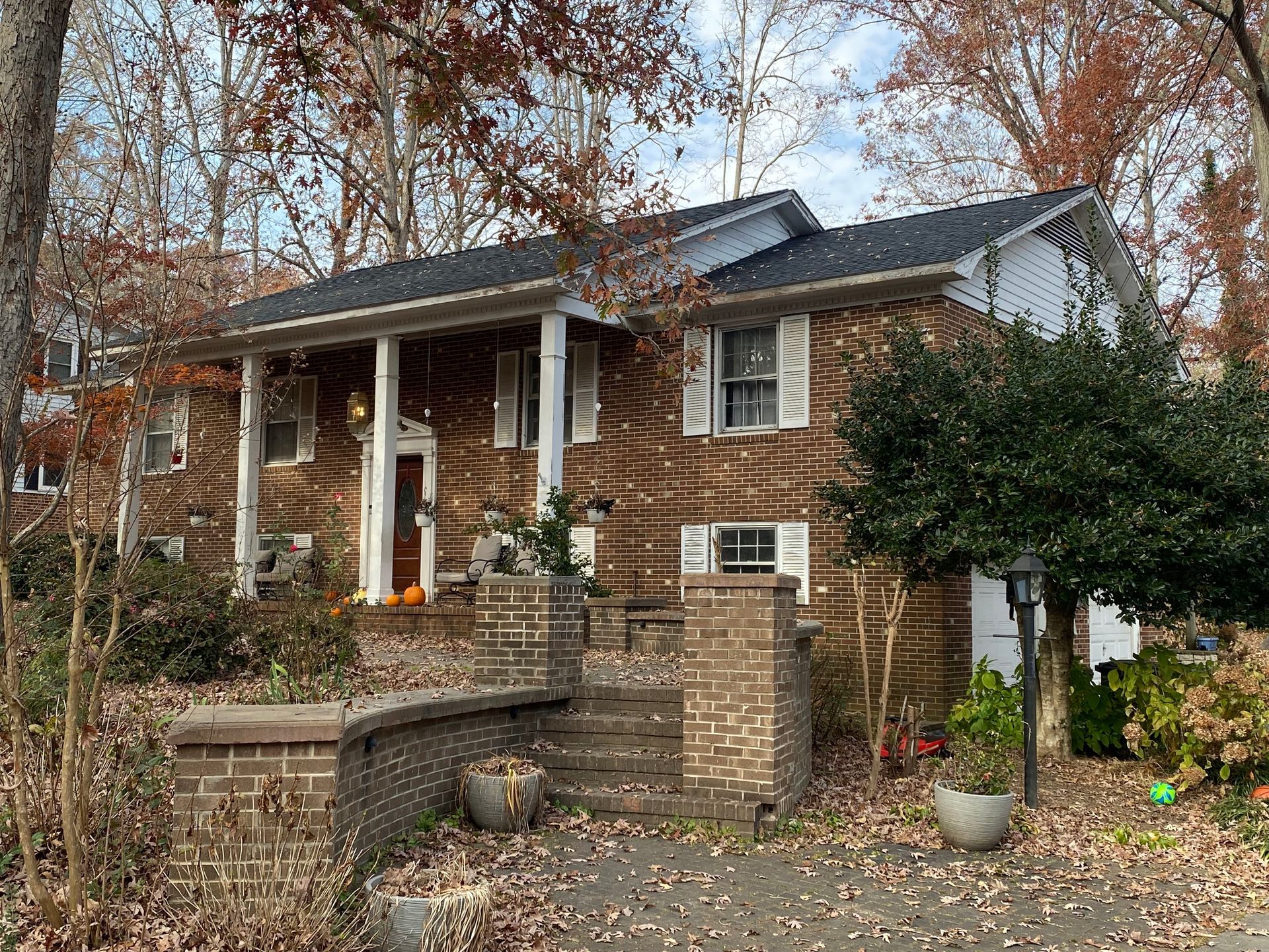 A two-story brick house with a white-columned front porch, surrounded by fall foliage and a brick wall with front steps.