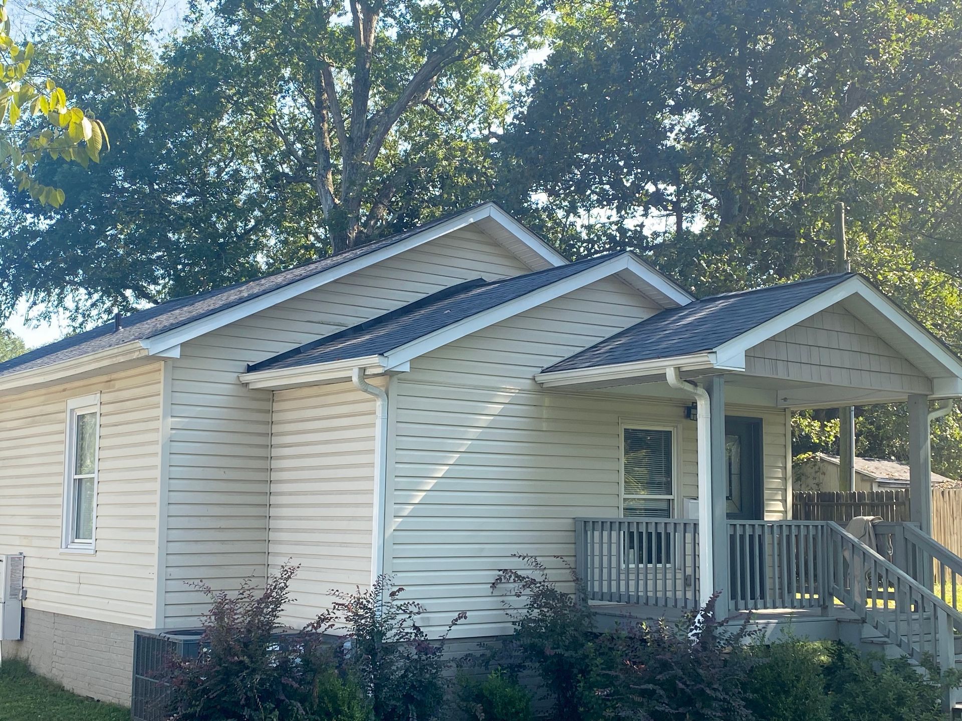 A one-story house with light-colored siding, a dark metal roof, and a front porch, situated among large green trees.