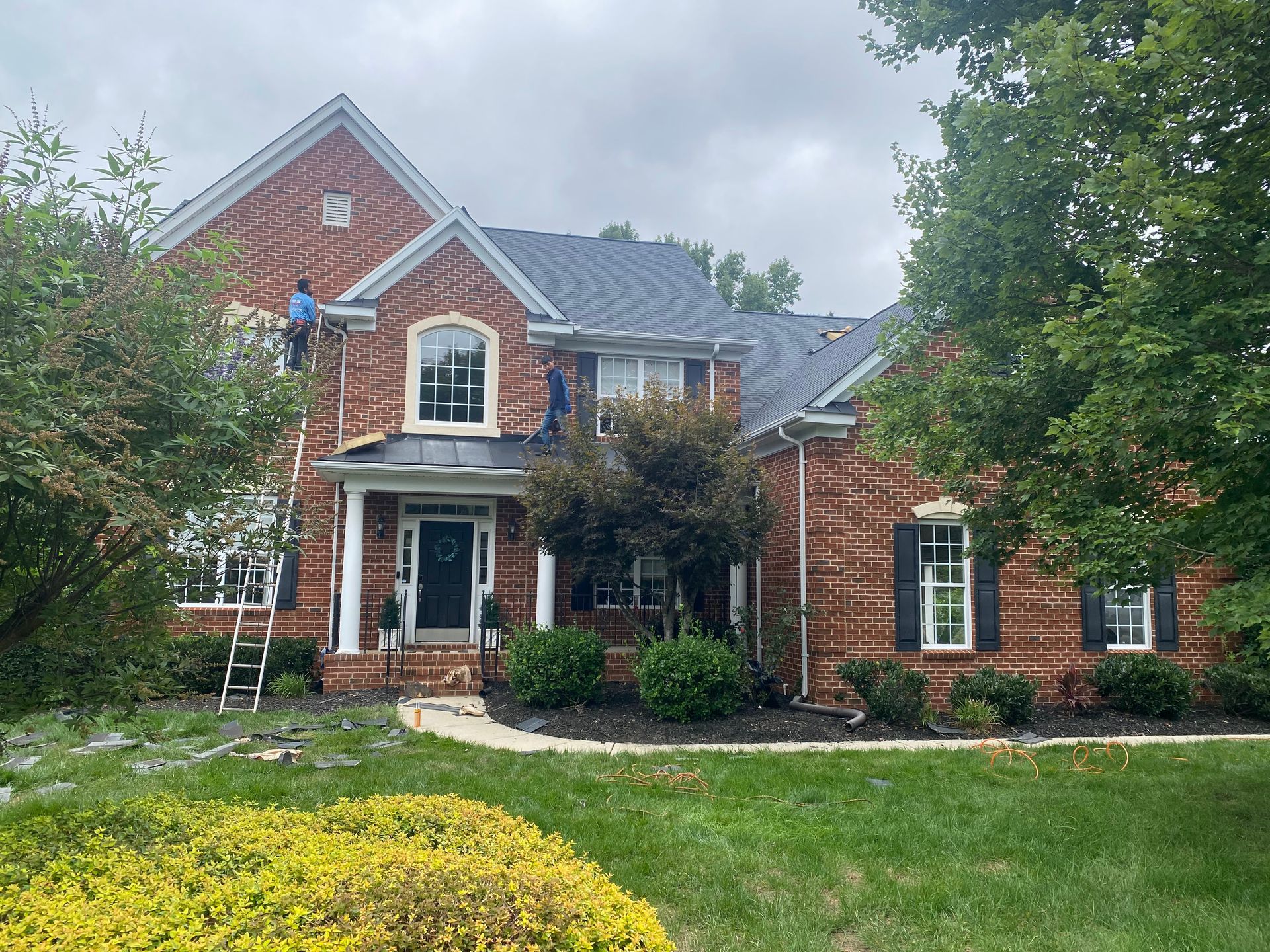 Workers on ladders perform roof repairs on a two-story brick home surrounded by trees and landscaping.