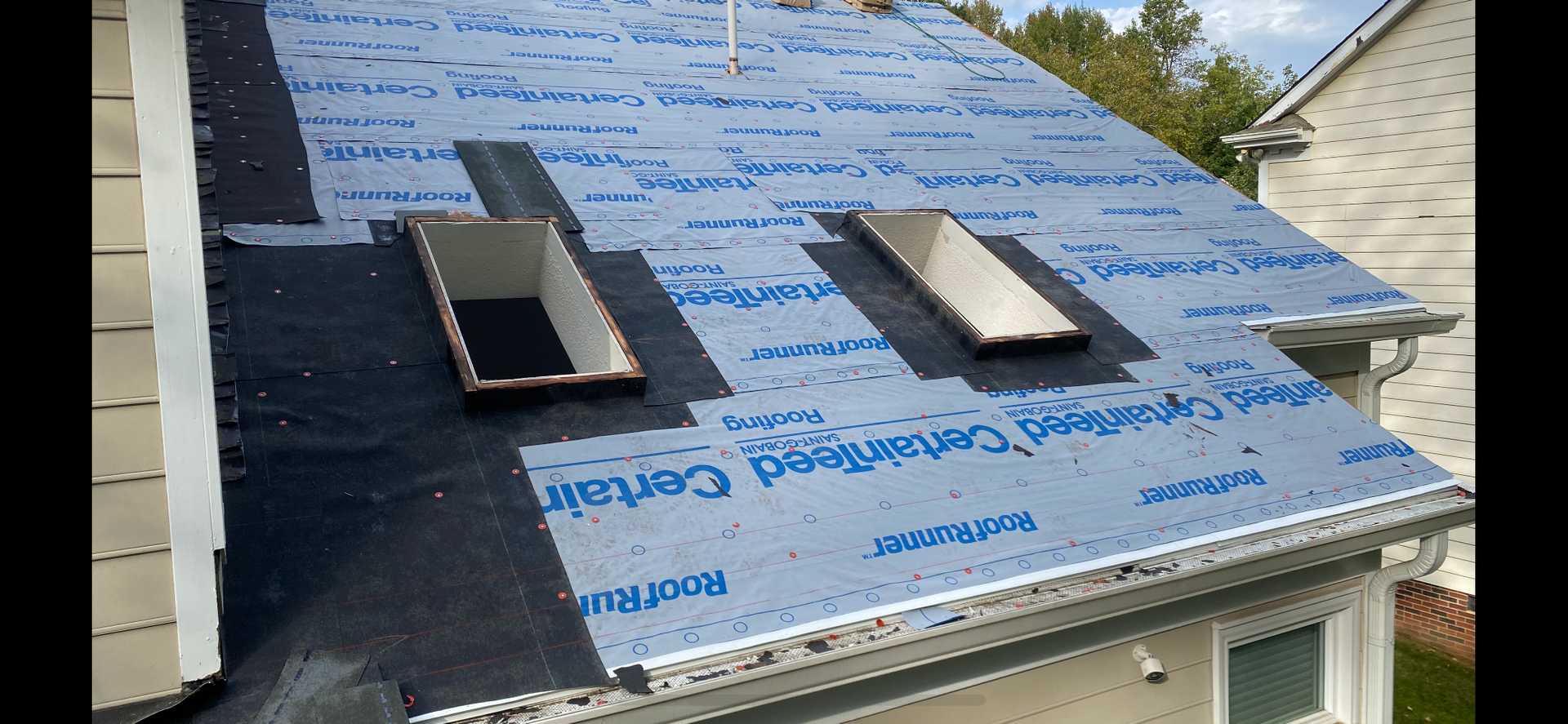 A residential roof under renovation, featuring two skylights installed over blue synthetic underlayment.
