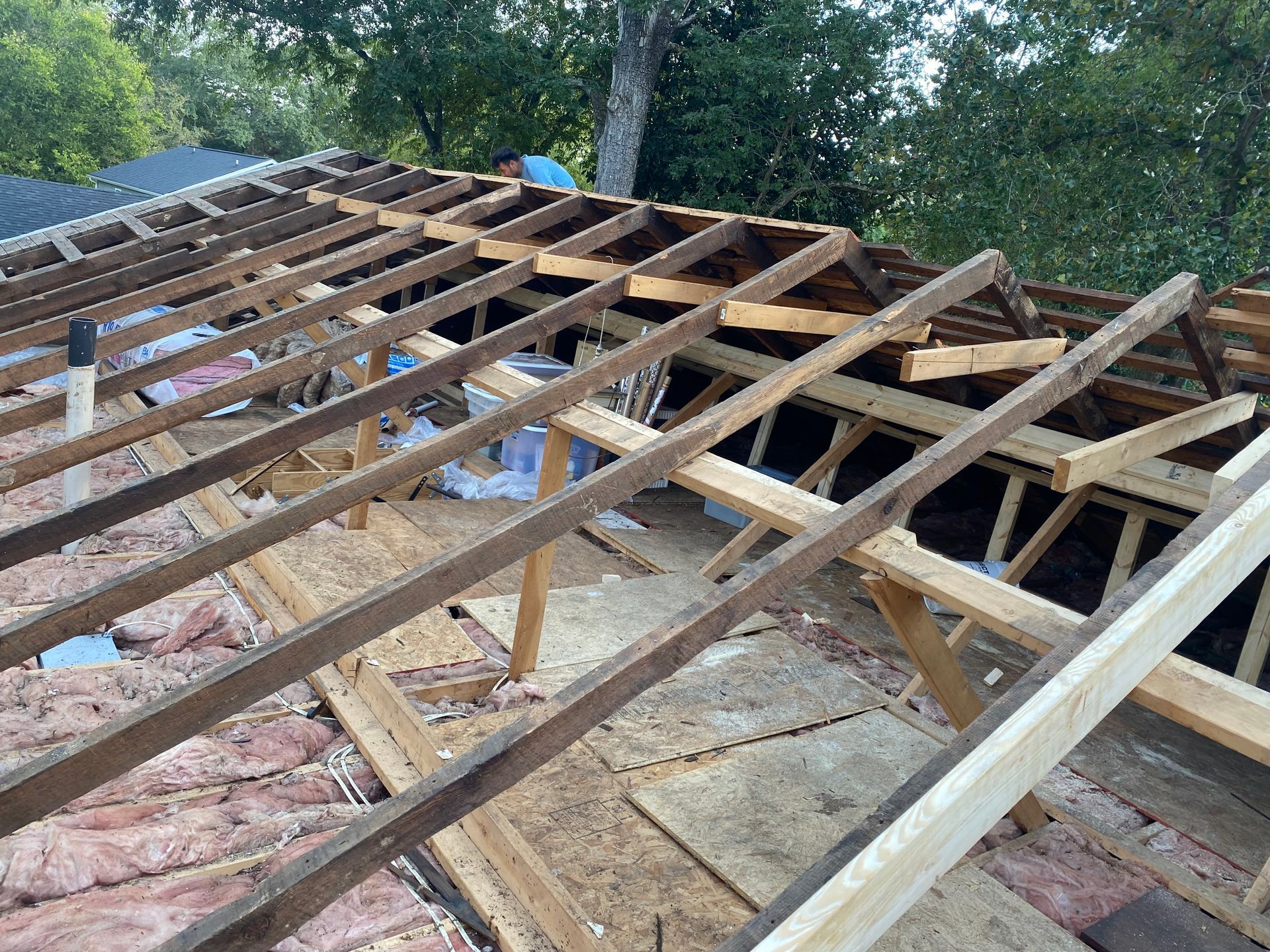 A roof undergoing repairs, showing exposed wooden rafters, insulation between the joists, and a worker in the background.