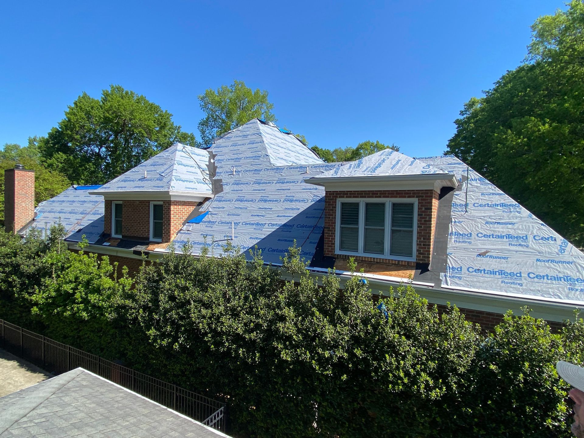 A roof under construction, covered in blue protective underlayment with two dormer windows, surrounded by green trees.