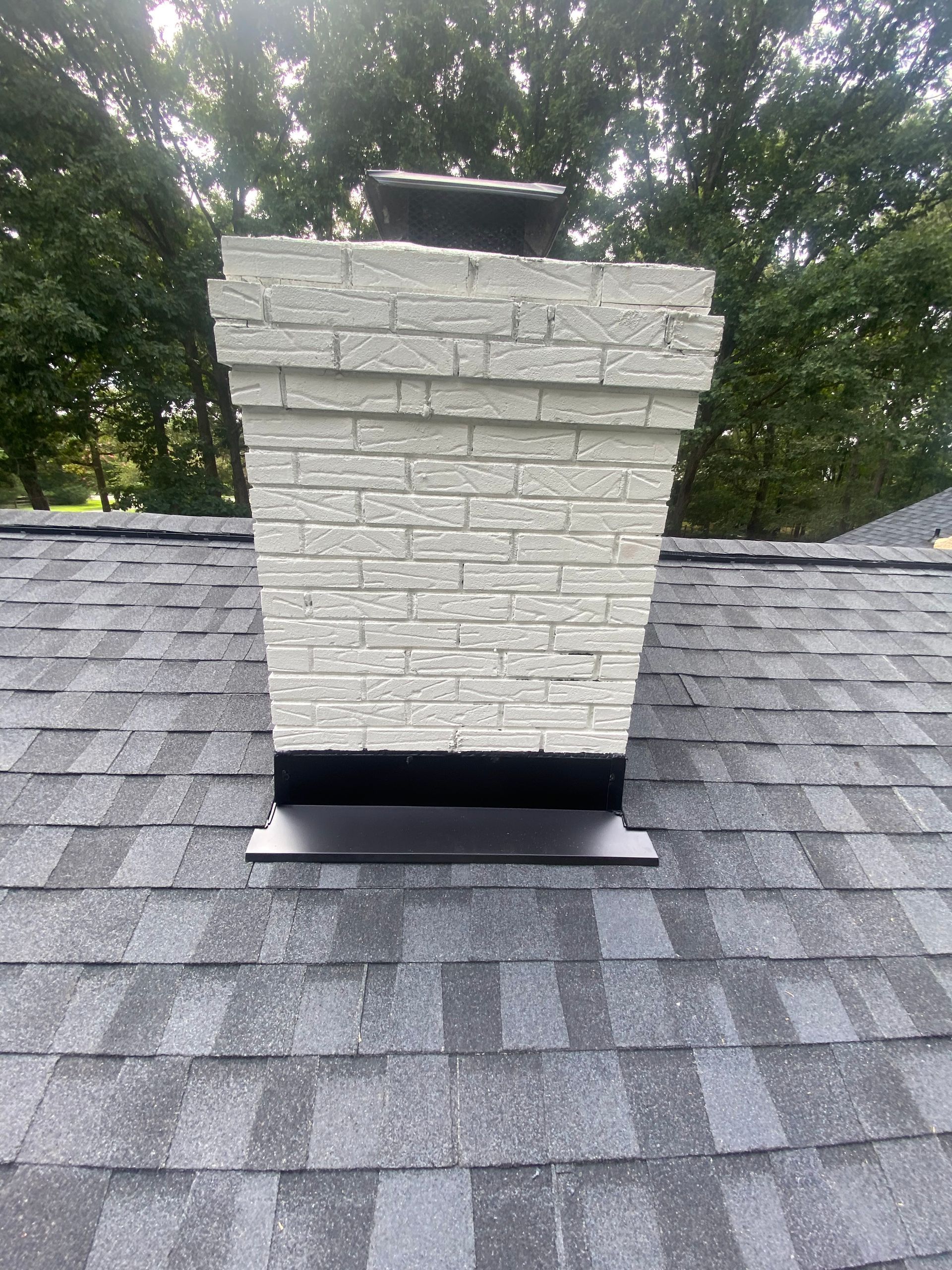 A white brick chimney with black flashing, set against a dark gray asphalt shingle roof and a background of green trees.