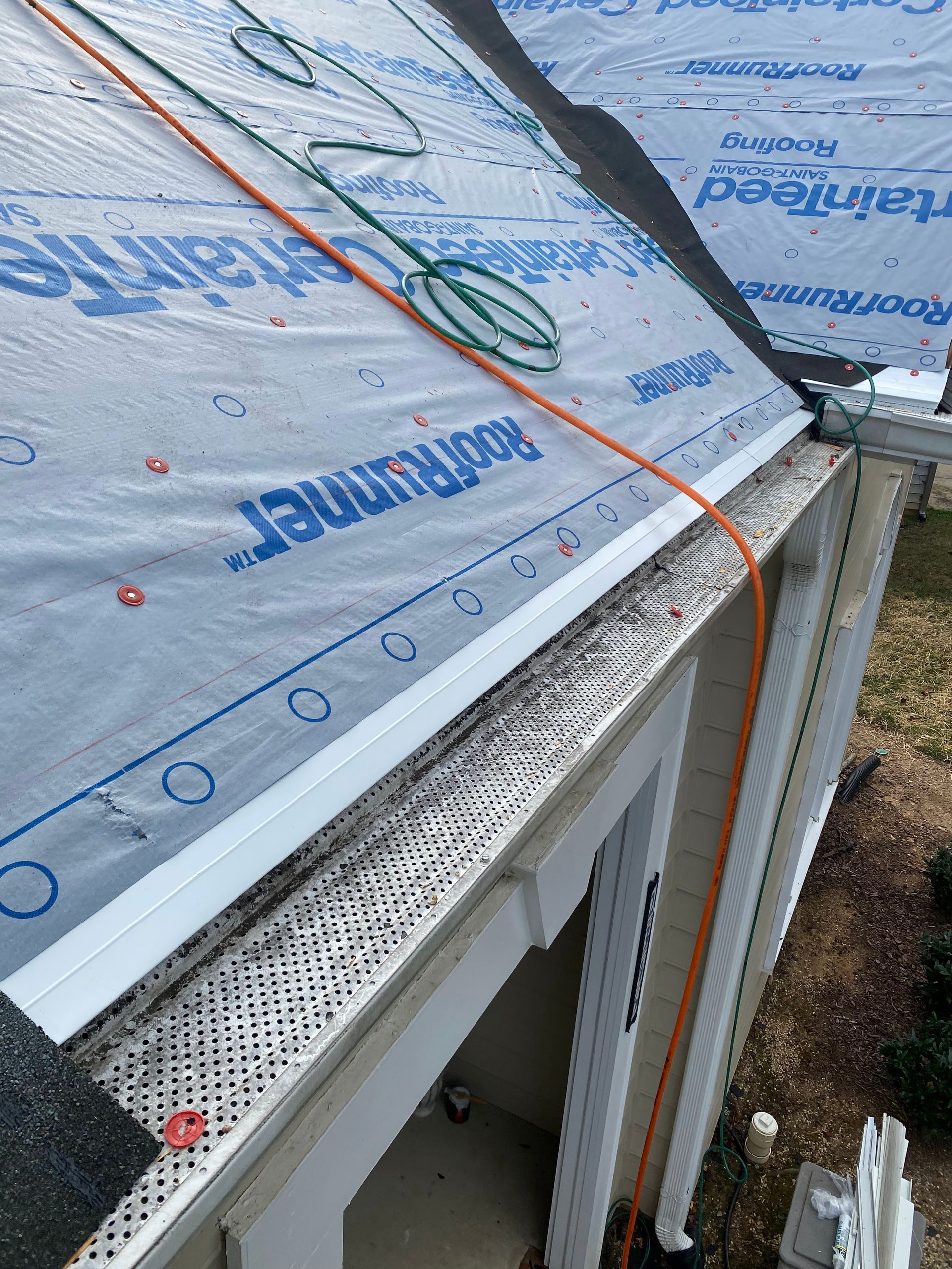 A high-angle view of a roof under construction, featuring blue synthetic underlayment over white fascia and gutters.