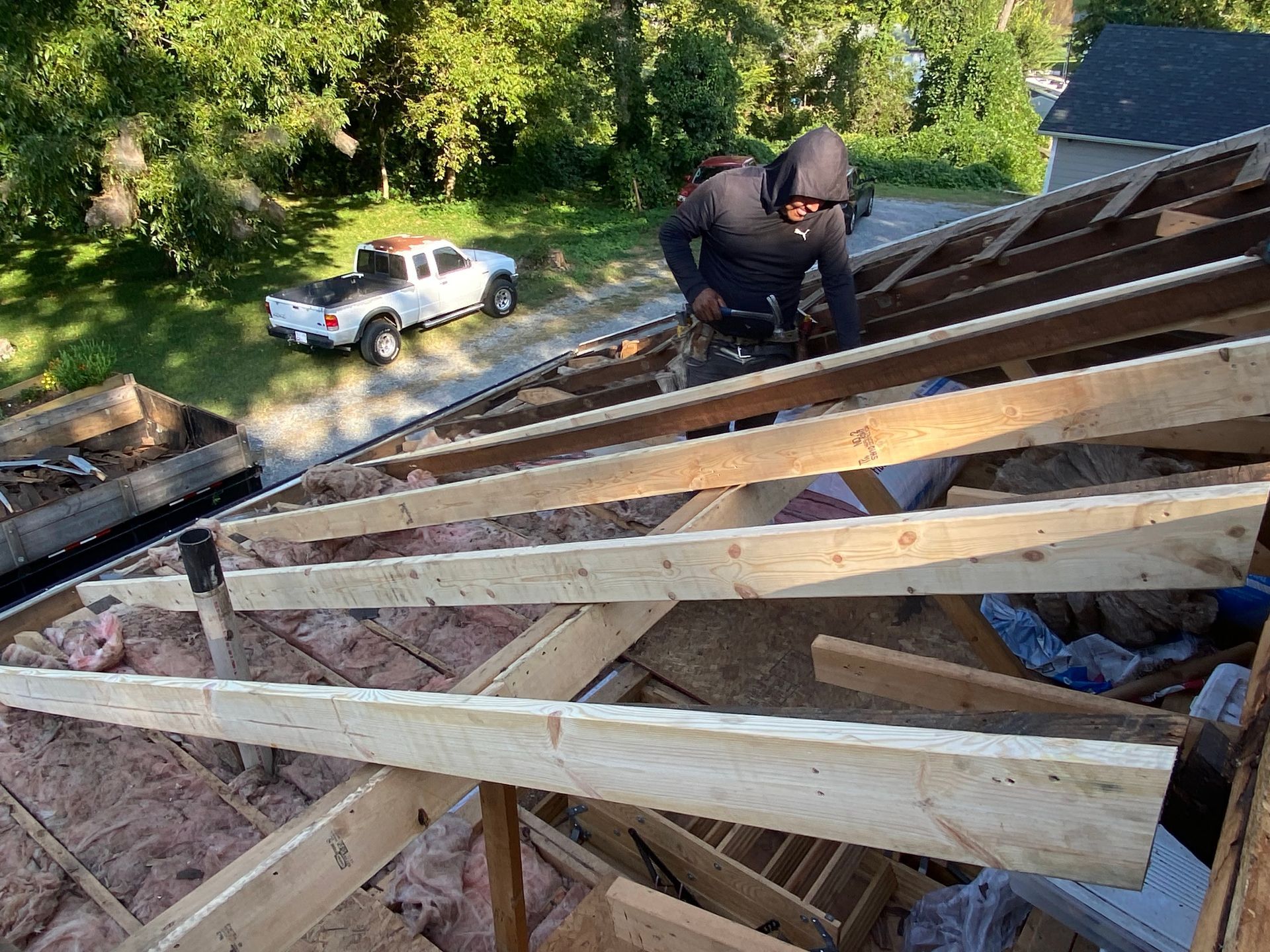 A worker in a hoodie installs wooden rafters on a roof frame over an attic with insulation, viewed from above.
