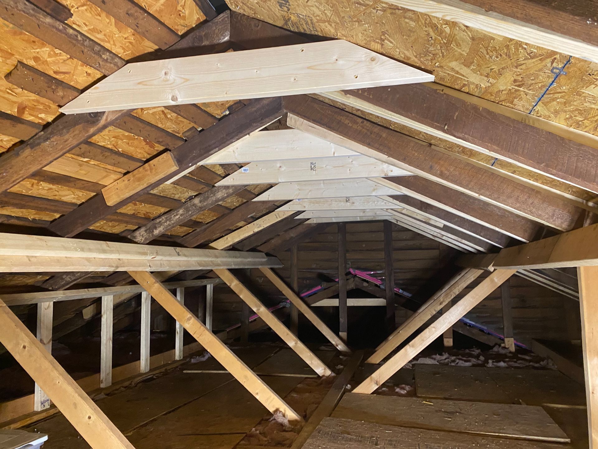 An attic interior showing exposed wooden roof rafters, trusses, and plywood flooring under a pitched roof.
