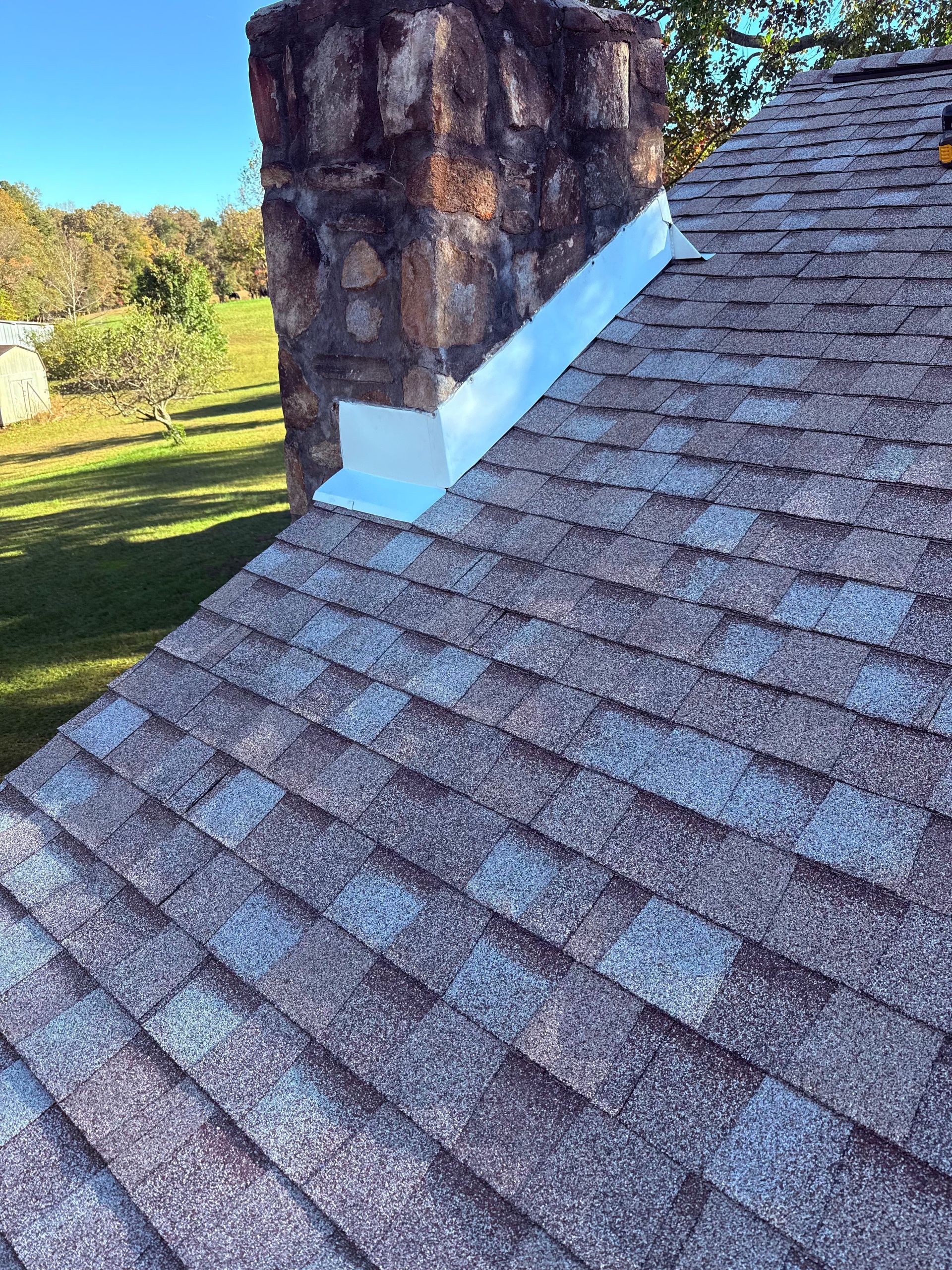 A stone chimney on a shingled roof with a bright white metal flashing seal at the base against the chimney stones.