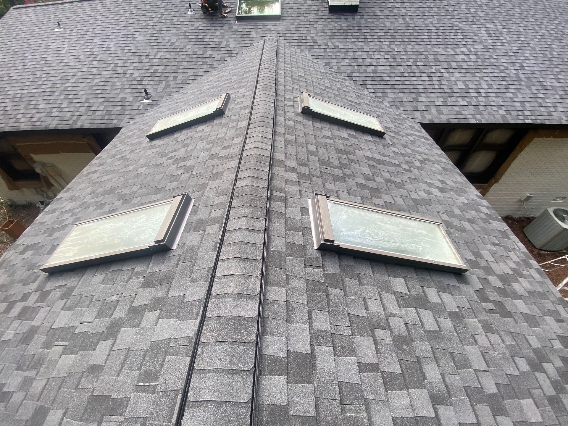 A gray shingled residential roof with four skylights, viewed from an elevated angle looking down the ridge.