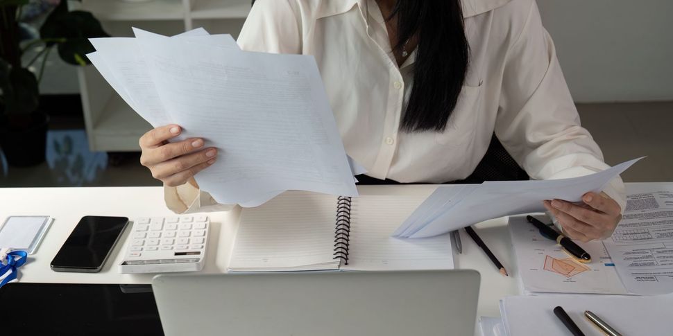 Person reviewing paperwork at a desk with a calculator, phone, and laptop. Person reviewing paperwork at a desk with a calculator, phone, and laptop.