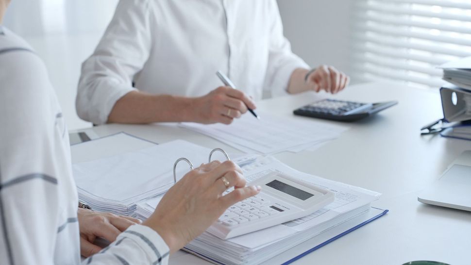 Two people calculating finances at a desk with papers, binders, and calculators in an office setting. Two people calculating finances at a desk with papers, binders, and calculators in an office setting.