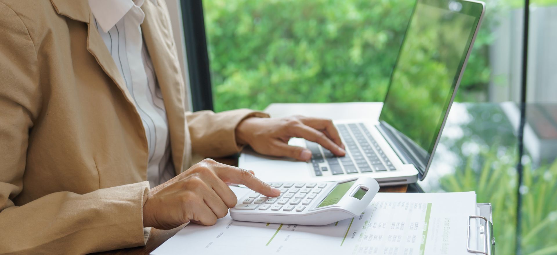Person using a calculator and laptop, working at a desk with paperwork, near a window with a green view. Person using a calculator and laptop, working at a desk with paperwork, near a window with a green view.