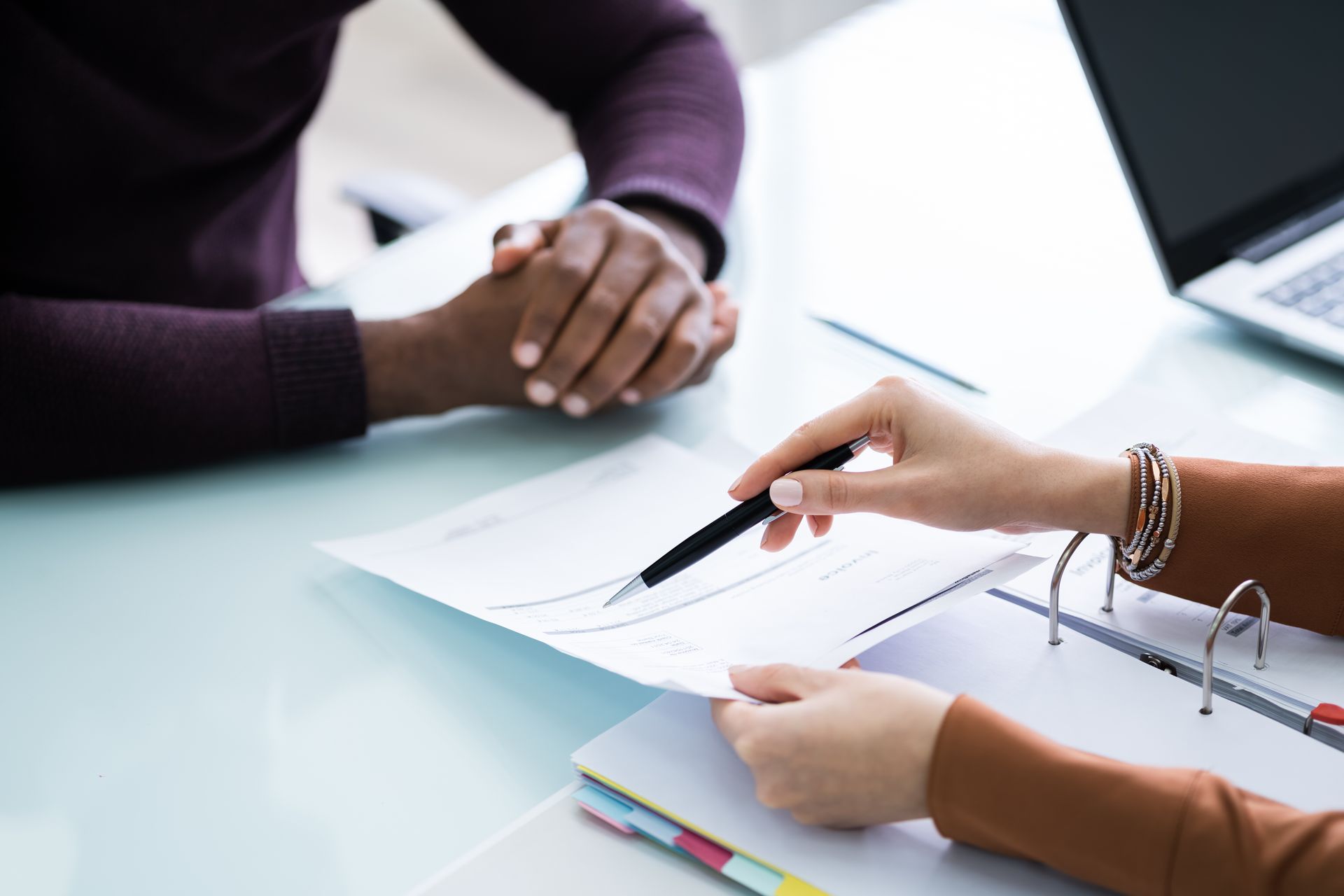 Person points at document; another person sits across, laptop in background. Person points at document; another person sits across, laptop in background.