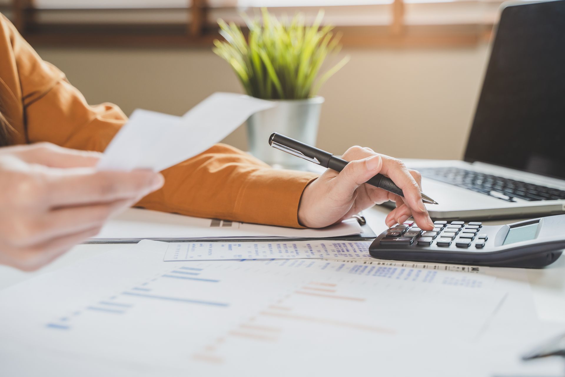 Person using a calculator, holding papers, and working on a laptop at a desk. Person using a calculator, holding papers, and working on a laptop at a desk.