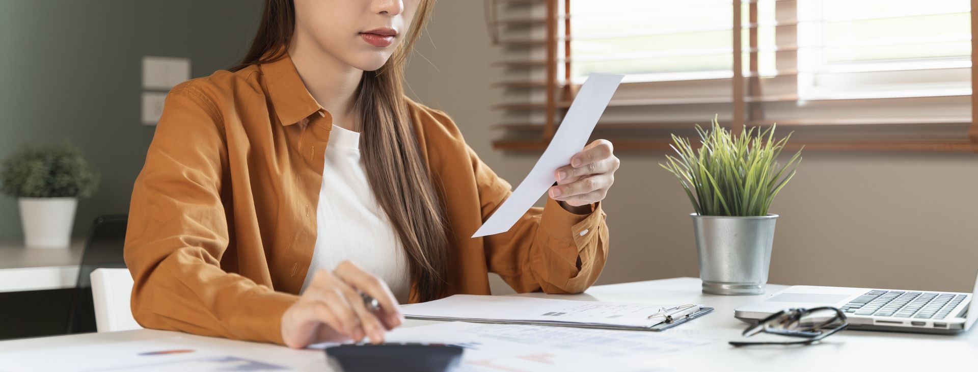 Woman reviews paperwork at a desk with calculator, laptop, and plant. Woman reviews paperwork at a desk with calculator, laptop, and plant.