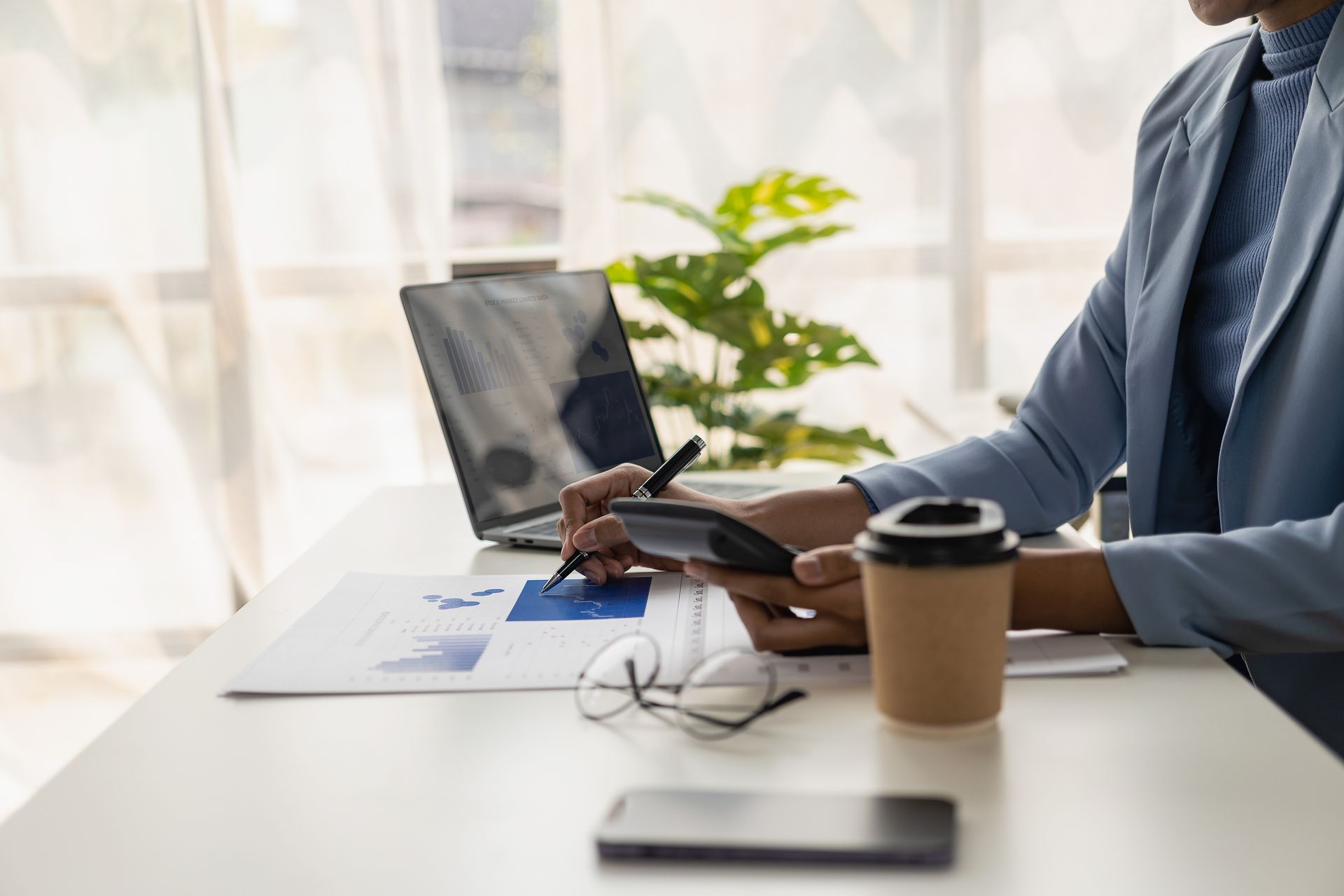 Person in blue blazer, calculating with a calculator, reviewing charts at a desk with a laptop and coffee. Person in blue blazer, calculating with a calculator, reviewing charts at a desk with a laptop and coffee.