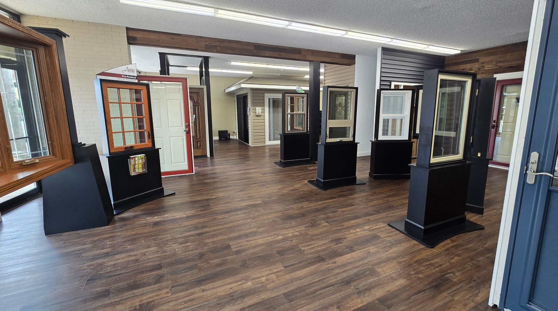 Showroom displaying various window and door styles, featuring wooden frames, dark floors, and overhead lighting.
