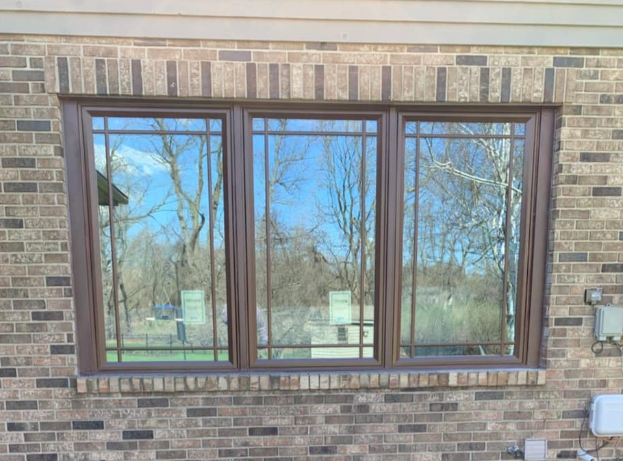 Brown-framed triple window with grid pattern set in brick wall; trees and sky reflected in glass.