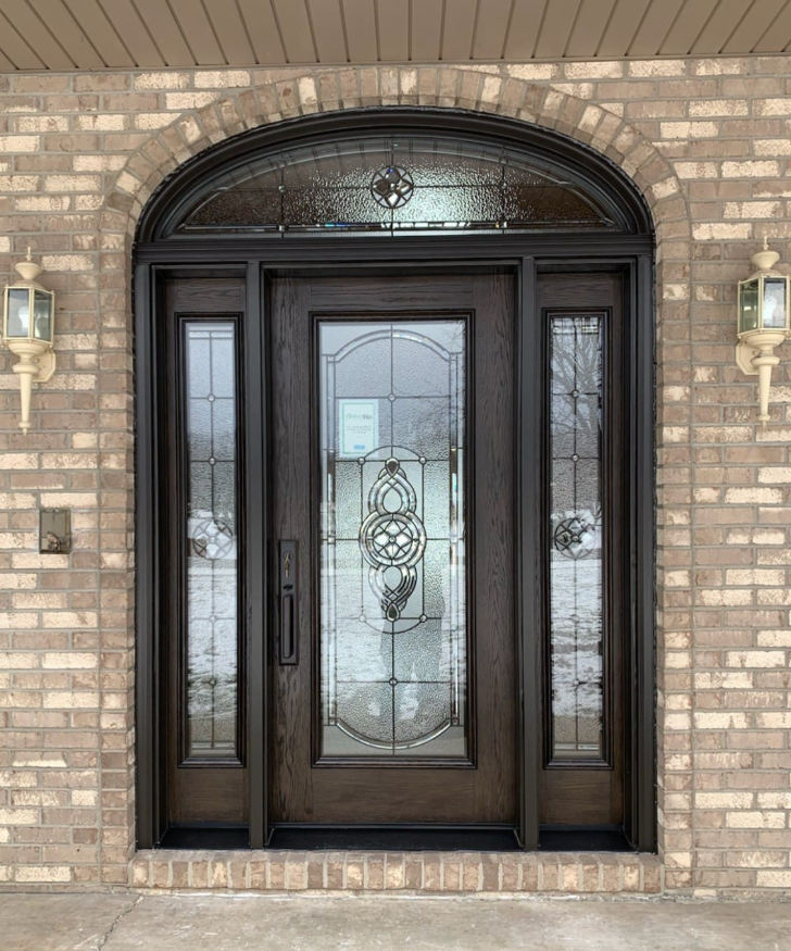 Brown front door with glass panels and arched transom, set in a brick wall, flanked by sconces.