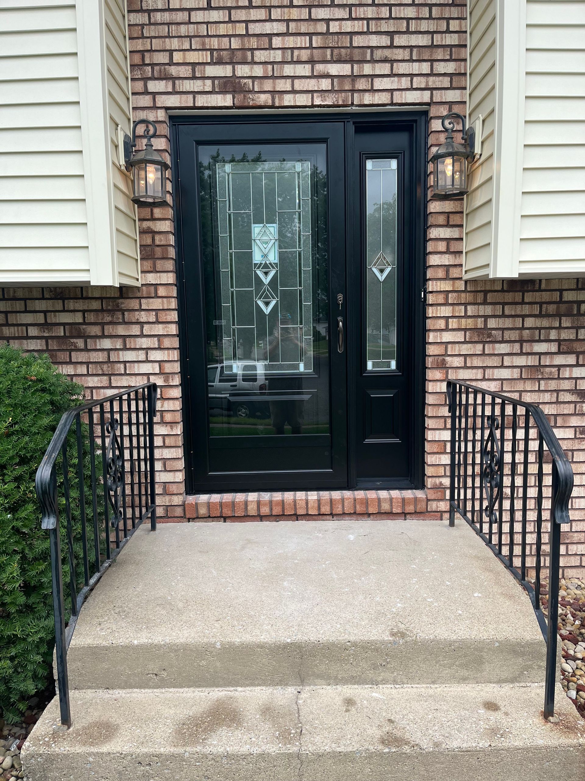 Black door with side panel and wrought iron railing leading to brick home.
