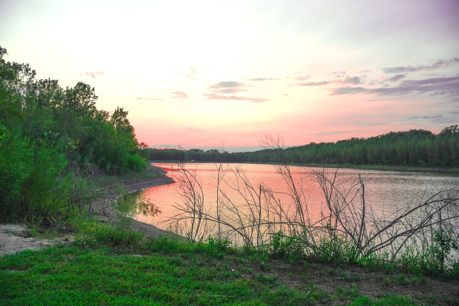 Sunset over a lake; orange and pink sky reflects on the water. Trees line the shore with green grass. Sunset over a calm river with pink sky, green banks, and silhouetted trees