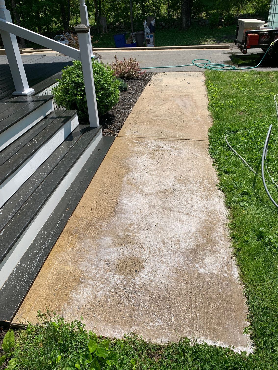 Concrete walkway with white residue leading from a porch to a paved area. Green grass and landscaping border the path.